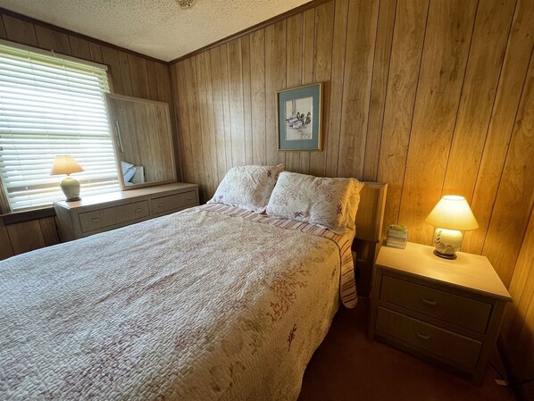 Une chambre à coucher comprenant un lit, des tables de chevet, un miroir, une fenêtre avec des stores et un tableau encadré accroché au mur.