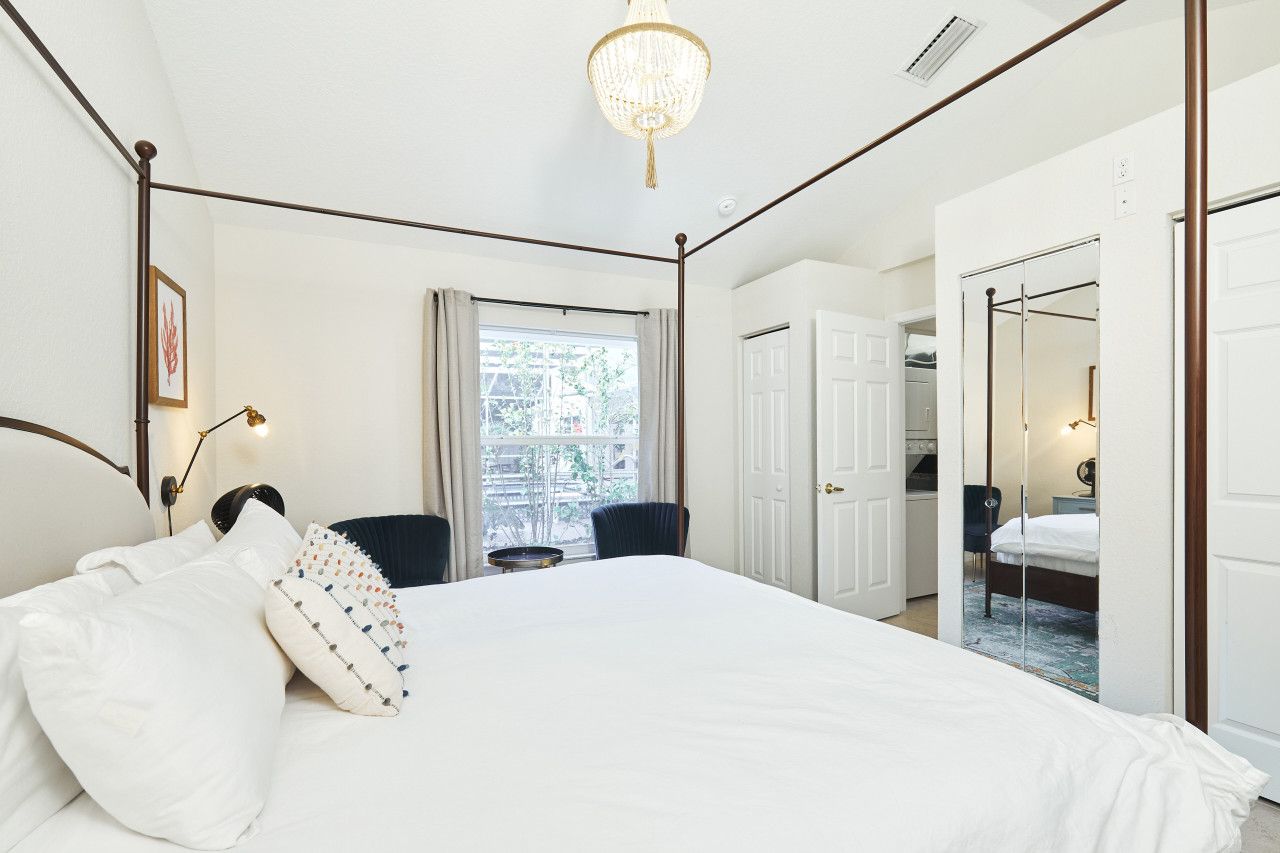 A bedroom with a four-poster bed, a chandelier, a window with curtains, and a door leading to another room.