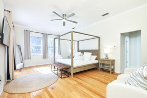 Una cama con dosel de estructura de madera, una mesita de noche con lámpara, un banco y un ventanal con cortinas.