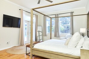 A four-poster bed with a wooden frame, a flat-screen TV mounted on the wall, a bedside table with a lamp, and a view of the outdoors through large windows. - The Oasis Retreat +Pool +Game Room +Basketball + Massive Yard +Near Beach & Golf (Largo)