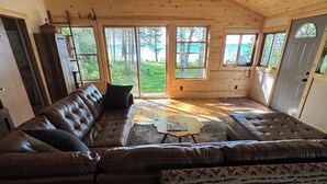Ein Holzblockhaus-Interieur mit einem großen, im L-Winkel angeordneten Ecksofa, einem gemusterten Teppich und großen Fenstern, die einen Blick ins Freie ermöglichen.
