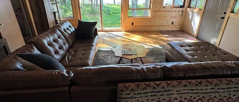A wooden cabin interior with a large L-shaped sectional sofa, a patterned rug, and large windows offering a view of the outdoors.
