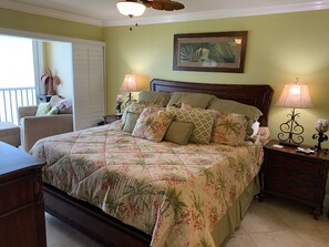 Quarto com colcha florida, cabeceira de madeira, duas luminárias de mesa, mesa de cabeceira, uma cadeira e janela com persianas.