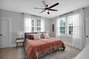A bedroom with a bed, a nightstand, a ceiling fan, and large windows with white curtains.