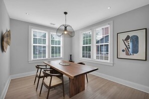 A dining area with a wooden table and chairs, large windows with blinds, a hanging light fixture, and a framed abstract artwork on the wall.