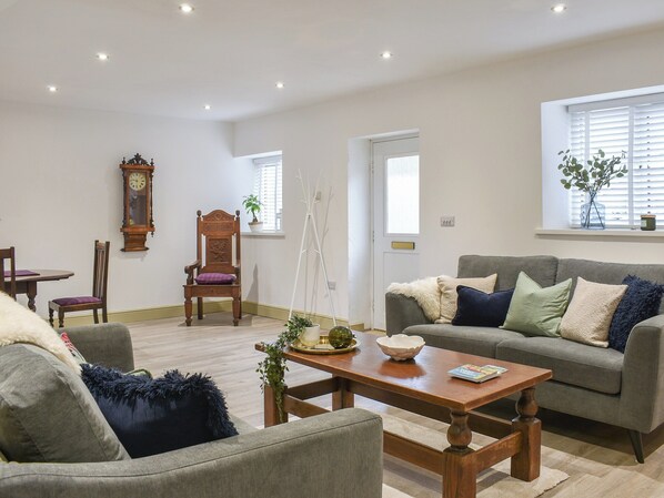Un salon avec un canapé gris, une table basse en bois, une chaise en bois, une horloge au mur et une fenêtre avec des stores.