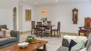 A living room with a sofa, a coffee table, a dining table with chairs, a clock, and a stone wall. - 2 bedroom accommodation in Abererch (Abererch)