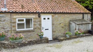 A stone cottage with a tiled roof, a white door, and a window. There are flower pots with pink and white flowers on the ground in front. - 1 bedroom accommodation in Gillamoor, near Kikbymoorside (Gillamoor, near Kikbymoorside)