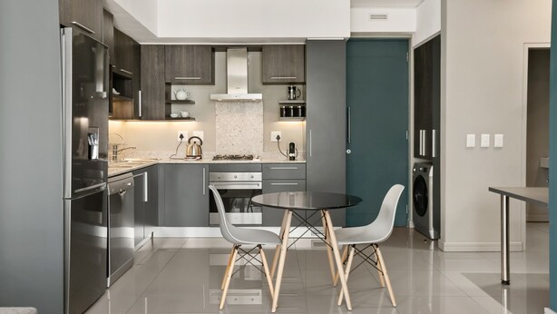 Een moderne keuken met roestvrijstalen apparatuur, grijze keukenkastjes, een ronde eettafel met twee stoelen en een accentmuur in turquoise.