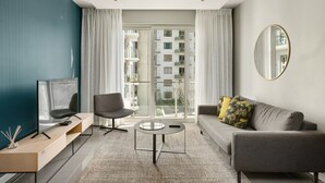 Een moderne woonkamer met een flatscreen televisie, een grijze bank, een ronde salontafel en een groot raam met gordijnen.