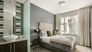 Een moderne slaapkamer met een groot bed, een nachtkastje met een lamp, een badkamer met een wastafel en spiegel, en een raam met gordijnen.