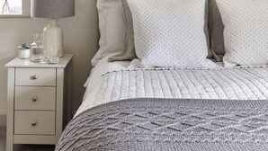 Una cama bien hecha con un edredón acolchado, una lámpara de noche y una mesita con un florero y vasos.
