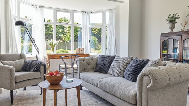 Un salón con sofá, sillón, mesa de centro de madera y vistas a la vegetación a través de amplias ventanas.