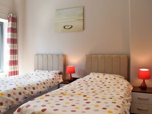 A hotel room with two beds, each with a polka dot bedspread, a red lamp on each nightstand, a wall-mounted heart-shaped decoration, and a window with striped curtains.
