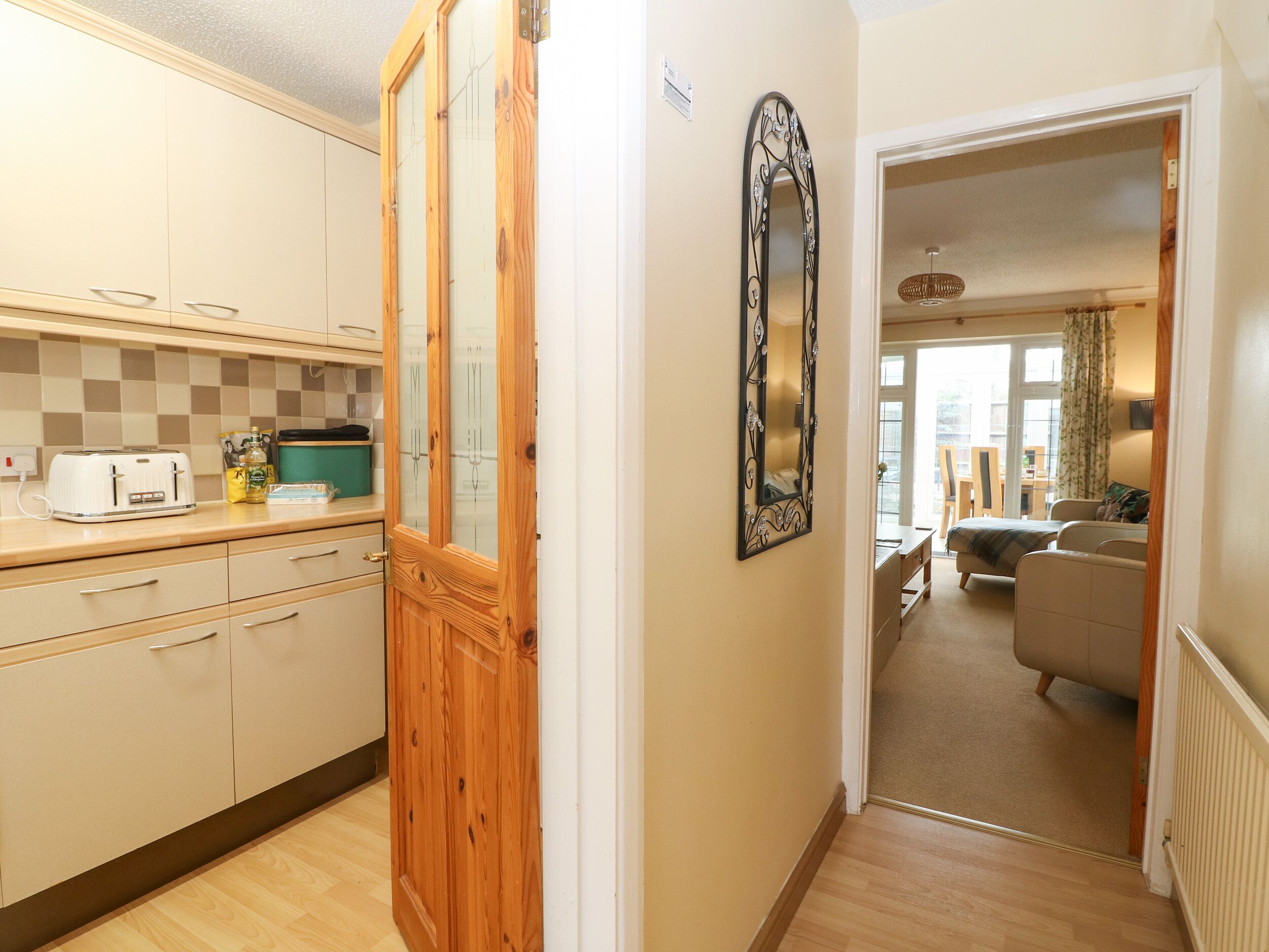 Eine Küche mit Holzausstattung, einem Toaster und einem grünen Behälter. An der Wand befindet sich ein Spiegel und eine Tür zu einem Wohnbereich mit Sofa und Tisch.