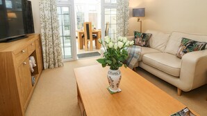 A living room with a television, a wooden coffee table with a vase of flowers, a beige sofa with decorative pillows, and a dining area visible through glass doors. - Maple Cottage (Broadway)