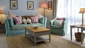 A living room with a sofa, armchair, coffee table, and two lamps. There are framed pictures on the wall and a window with sheer curtains. - The Butchers Cottage (Edzell, near Brechin)