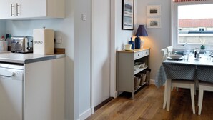 A kitchen with white cabinets, a toaster, a bread box, a lamp, and a dining table set for two. - The Butchers Cottage (Edzell, near Brechin)