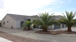 A two-story building with a sloped roof, a driveway, and several palm trees in the front yard. - Free Heated Pool, RV Garage, Hot Tub, Music, Views (Lake Havasu City)