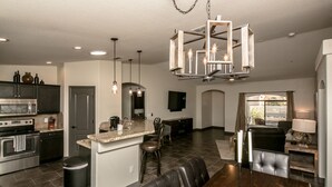 A modern kitchen with a central island, a dining area with a wooden table and chairs, and a living area with a sofa and a TV. - Free Heated Pool, RV Garage, Hot Tub, Music, Views (Lake Havasu City)