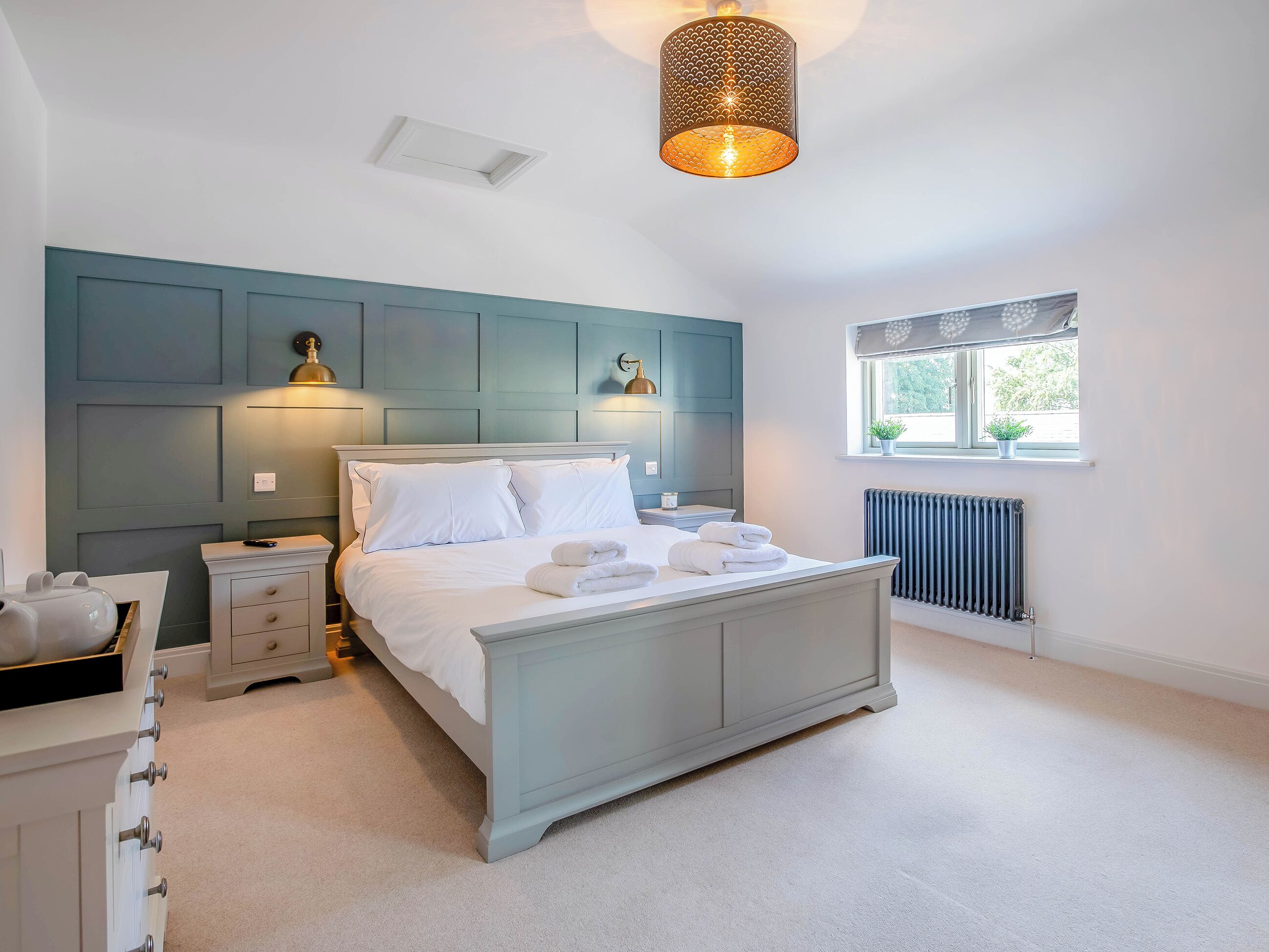 A bedroom with a bed, bedside tables, a radiator, a window with plants, and a hanging light fixture.