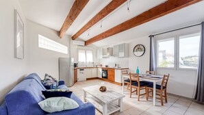 A modern living room with a blue sofa, a wooden coffee table, and a dining area with a table and chairs. A kitchen with white cabinets and a window is visible in the background. - Le Pescadou - Terrasse vue mer (Marseille)