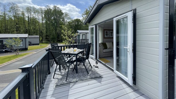 A deck with a table and chairs, a sliding glass door leading to a living area, and a view of a road and trees. - Luxurious and Cosy Holiday Lodge in the New Forest National Park (Landford)