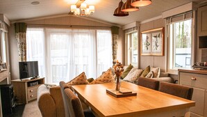 A cozy living room with a wooden dining table, comfortable seating, a television, and a kitchen area. - Luxurious and Cosy Holiday Lodge in the New Forest National Park (Landford)