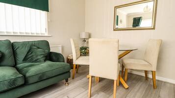 Una habitación con sofá verde, una mesa de comedor con sillas, un espejo en la pared y una ventana con persianas.
