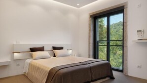 A modern bedroom with a large bed, a window with a view of greenery, and a thermostat on the wall.