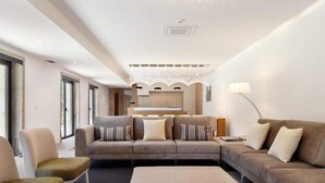 A modern living room with a sectional sofa, a small round table, and a bench. There is a kitchen area in the background with a large window.