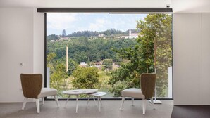 A modern room with a large window, two wicker chairs, a small round table, and a view of a green landscape with buildings.
