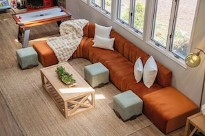 Un salon moderne avec un canapé d’angle, des poufs, une table basse en bois et de grandes fenêtres.