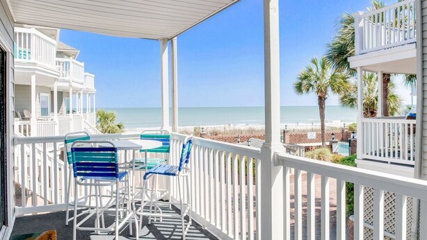 A covered balcony with white railings, a table, and blue chairs overlooking a beach.