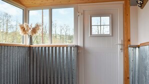 Ett rum med ett träpanelat tak, en vit dörr med ett litet fönster, ett fönster med metallram, en vägg av metall, en säng med ett rutigt sängtäcke och en dörrmatta.