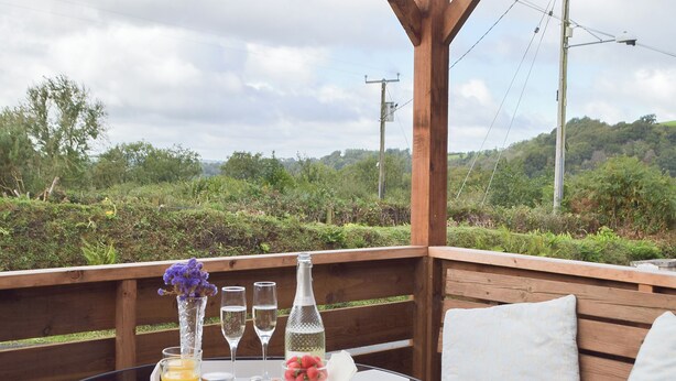 A wooden deck with a round table set for a meal, featuring a bottle, glasses, and a fruit bowl.