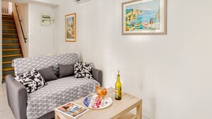 A living room with a grey patterned sofa, a wooden coffee table with a bottle and glasses, a framed painting, and a staircase.