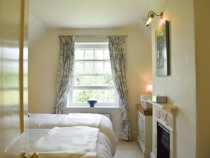 A bedroom with two beds, a window with curtains, a bedside table with a lamp, and a fireplace.