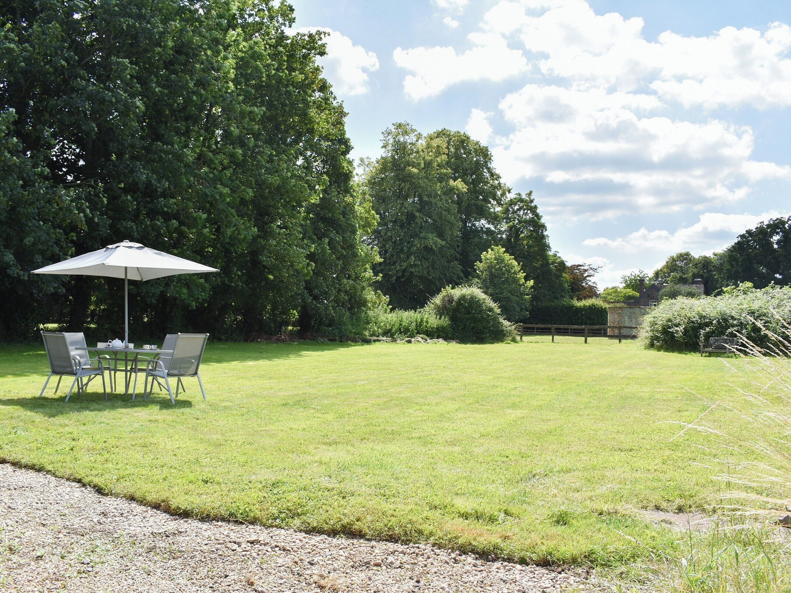 A grassy outdoor area with a table and chairs under a large umbrella, surrounded by trees and a stone wall.