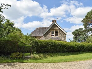 A stone house with a gabled roof, surrounded by lush greenery and a gravel driveway.