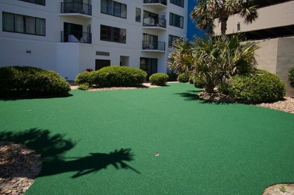 A courtyard with artificial green turf, surrounded by neatly trimmed bushes and a palm tree.