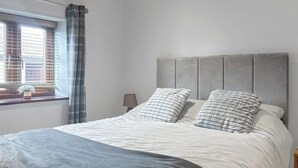 A neatly made bed with a grey headboard and two pillows, a bedside table with a lamp, and a window with blinds and a curtain. - 2 bedroom accommodation in Catsham, near Glastonbury (Catsham, near Glastonbury)