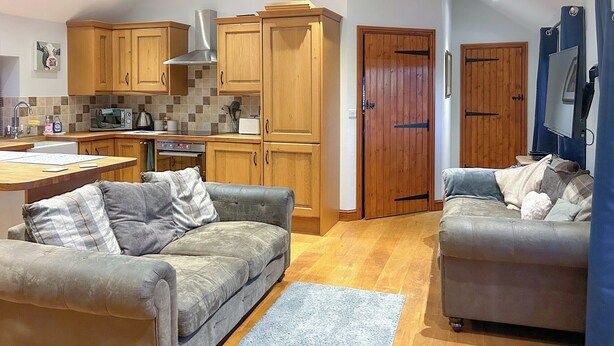 A modern kitchen with wooden cabinets and a built-in microwave. There are two grey sofas with cushions, a wooden dining table, and a blue rug on the floor. - 2 bedroom accommodation in Catsham, near Glastonbury (Catsham, near Glastonbury)