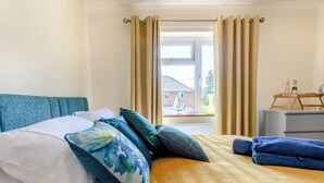 Una cama bien tendida con un cojín estampado, un cabecero azul y vista a una casa a través de una ventana con cortinas color beige.