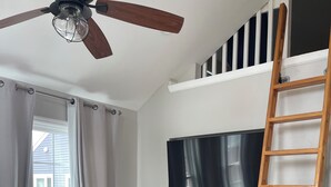 Un coin repas avec une table et des chaises en bois, un ventilateur de plafond, un téléviseur fixé au mur et une échelle en bois.