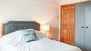 Un lit bien fait, doté d’une tête de lit rembourrée de couleur grise, d’une lampe de chevet et d’une armoire en bois à deux portes.