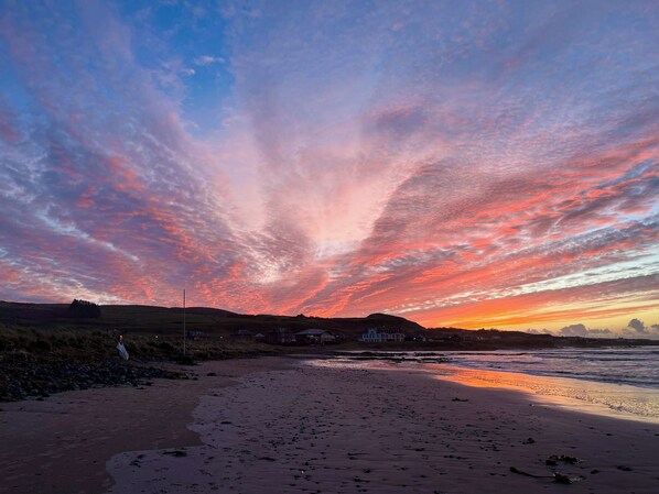 Una playa con un atardecer, casas al fondo y un cielo despejado.