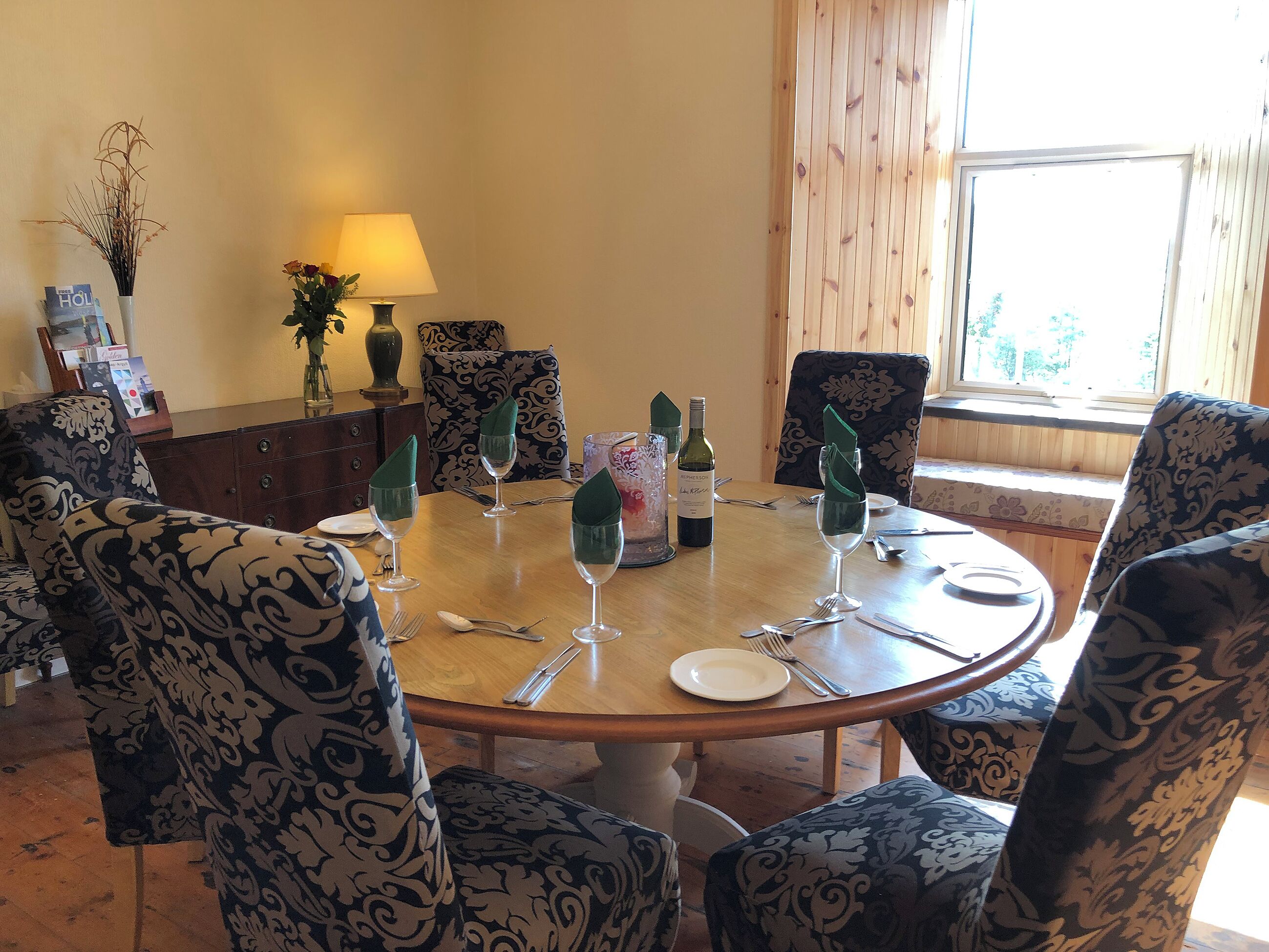 Un comedor con una mesa redonda, sillas estampadas, una botella de vino y un florero con plantas secas.