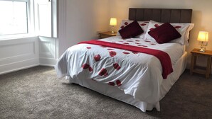 Un lit bien fait, recouvert d’une couvre-lit à motifs floraux blancs et rouges, avec deux oreillers rouges et un plaid rouge. Deux lampes de chevet sont posées sur des tables de chevet en bois.