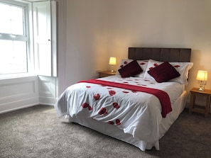 Una cama bien hecha con colcha blanca y estampada con motivos florales, dos almohadas rojas y una manta roja. Hay dos lámparas de noche sobre mesitas de madera.